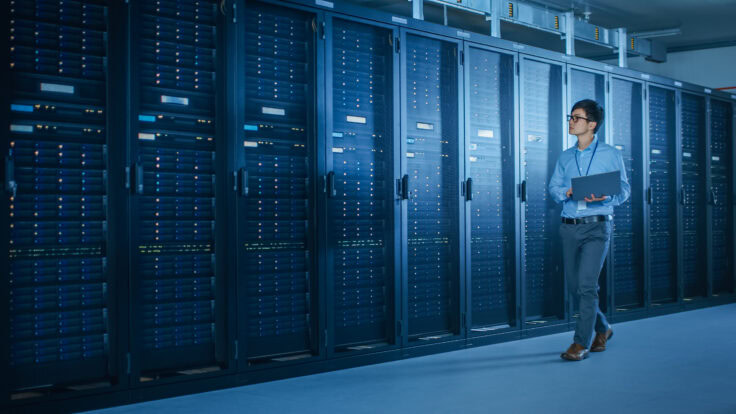 In Data Center: Male It Technician Running Maintenance Programme On A Laptop, Controls Operational Server Rack Optimal Functioning. Modern High Tech Telecommunications Operational Super Computer.
